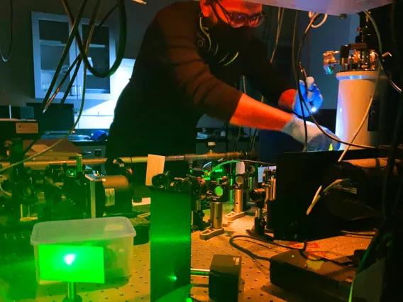Student working at table covered in optics with green laser light in foreground
