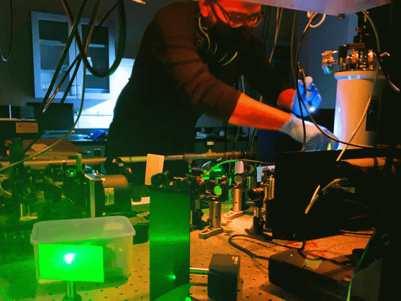 Student working at table covered in optics with green laser light in foreground